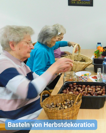 Seniorinnen sitzen gemeinsam an einem Tisch. Vor ihnen sind Körbe mit Naturmaterialien, unter anderem Kastanien. Bildbeschriftung: Basteln von Herbstdekoration