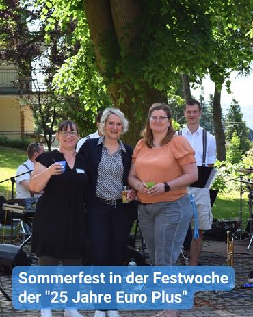 Das Bild zeigt eine Gruppe von vier Personen, die an einem warmen Sommertag draußen stehen. Drei Frauen befinden sich im Vordergrund, während ein Mann im Hintergrund steht.  Die Frau links trägt ein schwarzes Kleid und eine Brille, während sie eine blaue Tasse hält. Die mittlere Frau hat blonde Haare und trägt eine gemusterte Bluse, kombiniert mit einem dunklen Blazer und dunklen Hosen. Die Frau rechts trägt ein orangefarbenes Oberteil sowie graue Hosen und hält eine kleine grüne Tasse. Der im Hintergrund stehende Mann trägt ein weißes Hemd mit Hosenträgern, leichte Shorts und hält ein Notenständer.  Die Gruppe befindet sich in der Nähe eines großen Baumes und musikalischer Ausrüstung, was darauf hindeutet, dass sie möglicherweise an einer Open-Air-Veranstaltung oder einem Festival teilnehmen. Ein blaues Banner mit dem Text „Sommerfest in der Festwoche der »25 Jahre Euro Plus«” ist am unteren Rand des Bildes sichtbar.