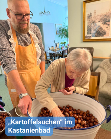 Betreuer und Seniorin stehen vor einer großen Schüssel voller Kastanien. Die Seniorin greift hinein. Bildbeschriftung: „Kartoffelsuchen“ im Kastanienbad