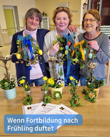 Drei Frauen stehen in einem Raum und lächeln in die Kamera. Sie halten frühlingshafte Bastelarbeiten, die kleine Arrangements aus Zweigen, Grünpflanzen und Ostereiern beinhalten. Diese Arrangements sind auf einem Tisch vor ihnen ausgestellt, zusammen mit weiteren kleinen dekorativen Gegenständen in Gläsern und Töpfen. Auf dem Tisch liegen zudem Zertifikate, die darauf hindeuten, dass die Frauen eine Art Kurs oder Schulung abgeschlossen haben.  Die Frauen sind lässig gekleidet. Die Dame ganz links trägt einen mauvefarbenen Pullover. Die Frau in der Mitte präsentiert ein türkisfarbenes Shirt. Die Frau ganz rechts trägt ein gestreiftes Langarmshirt. Der Hintergrund ist leicht verschwommen, zeigt jedoch einen hellen Raum mit sichtbaren Möbeln und Dekoration.  Am unteren Rand des Bildes befindet sich ein Spruch: "Wenn Fortbildung nach Frühling duftet." Der Text ist in einem blauen Banner abgedruckt.