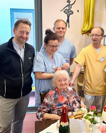 Eine Innenaufnahme auf Augenhöhe zeigt eine Gruppe von Menschen, die sich um eine ältere Frau versammelt haben, die an einem Tisch mit Torten und Champagner sitzt. Es handelt sich um den 102. Geburtstag dieser Dame.  Die ältere Frau nimmt eine zentrale Position ein, ist in ein blumiges Kleid gekleidet und blickt mit einem Lächeln direkt in die Kamera. Sie wird von vier jüngeren Erwachsenen umgeben, darunter zwei Männer und zwei Frauen. Ein Mann steht links von ihr, bekleidet mit einer dunkelgrauen Zip-up-Jacke und hellgrauen Hosen. Ein weiterer Mann steht leicht hinter ihr rechts, gekleidet in einem hellblauen Poloshirt. Die beiden Frauen stehen jeweils an den Seiten der sitzenden Dame; die zu ihrer linken trägt ein hellblaues Hemd, während die zu ihrer rechten ein senfgelbes Shirt anhat. Ein großer goldener Luftballon ist teilweise im Hintergrund sichtbar.  Die Szenerie ist deine Geburtstagsfeier in einer Wohn- oder Seniorenresidenz. Im Hintergrund ist eine Tür zu sehen, die zu einem weiteren Raum oder Flur führt. Die Wände sind hell gehalten, und es gibt eine Wanddekoration, die eine Ballerina vor einem schwarzen Hintergrund zeigt. Auf dem Tisch sind Geburtstagstorte, Teller, Gläser und eine Champagnerflasche dekorativ angerichtet. Insgesamt strahlt die Atmosphäre Wärme und Feierlichkeit aus.