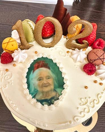 Eine festliche Torte, zum 102. Geburtstag, steht im Mittelpunkt dieser Nahaufnahme. Die Zahl "102" thront prominent auf der Torte, gestaltet aus goldfarbenem, glitzerndem Material. Um die Ziffern herum sind verschiedene bunte Dekorationen angerichtet: Erdbeeren, Himbeeren, Makronen und weitere Süßigkeiten. Zudem finden sich Schokoladenstücke und kleine, dekorierte Bonbons.  Die Torte selbst ist rund und weiß, verziert mit einem filigranen Randmuster. Ein zentrales Merkmal ist ein essbares Abbild einer älteren Dame,  dem 102-jährigen Geburtstagskind, gekleidet in einer dunklen Bluse. Ihr Porträt ist in einem ovalen Ausschnitt eingefasst, was das elegante Design der Torte unterstreicht.  Die Torte ruht auf einer goldfarbenen Tortenplatte, und ein kleines, teilweise sichtbares Bäckerei-Label ist unten rechts eingeklemmt. Der Hintergrund besteht aus einer verschwommenen, dunkelbraunen Fläche, die dazu beiträgt, die strahlende und farbenfrohe Torte hervorzuheben.