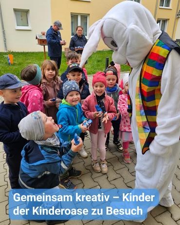 Auf Augenhöhe sieht man eine Gruppe von Kindern, die sich um ein großes, weißes Kostüm eines Osterhasen versammeln.  Die Kinder, eine Mischung aus Jungen und Mädchen im Grundschulalter, sind in bunte Winterkleidung gehüllt. Einige halten kleine Gegenstände in den Händen, möglicherweise Ostereier oder Süßigkeiten. Der Hase wirkt freundlich und interagiert mit den Kindern. Die Szenerie ist im Freien, vor einem Gebäude, das wie ein Appartementkomplex oder eine Kindertagesstätte aussieht. Im Hintergrund ist ein Erwachsener teilweise sichtbar, der eine dunkle Jacke trägt.  Die Szene strahlt eine helle und fröhliche Atmosphäre aus, suggestiv für eine Osterveranstaltung oder einen Besuch. Ein Banner am unteren Rand des Bildes trägt die Aufschrift „Gemeinsam kreativ - Kinder der Kinderroase zu Besuch.“