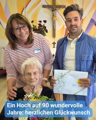 Foto einer 90-jährigen Seniorin, welcher von einer freundlichen Frau und einem Herrn gratuliert wird. Im Hintergrund ist eine Kapelle zu sehen. Bildunterschrift: “Ein Hoch auf 90 wundervolle Jahre: herzlichen Glückwunsch”