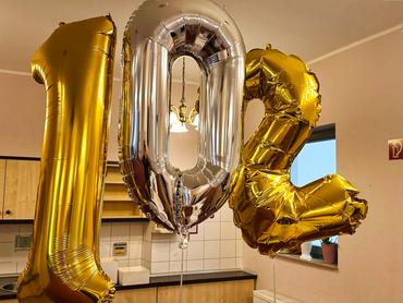 Nahaufnahme von drei großen Folienballons, die zusammen die Zahl "102" bilden.  Der Ballon, der die "1" darstellt, ist goldfarben, während der "0" silber ist und die "2" ebenfalls goldfarben. Die Ballons glänzen und reflektieren das Licht.  Die drei Ballons schweben vor einer Küchenzeile. Teile einer weißen Fliesenwand, hellfarbige Schränke und ein Abschnitt einer Arbeitsplatte sind sichtbar. Im Hintergrund hängt auch eine Art Kronleuchter. Die Gesamtbeleuchtung in der Szene ist sanft und warm.