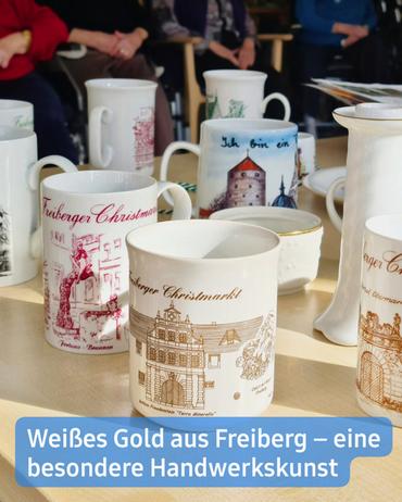 Eine hochwinkelige Nahaufnahme zeigt mehrere Tassen, die auf einer hellen Holzoberfläche stehen. Die meisten Tassen sind weiß und mit goldenen Verzierungen sowie Schriftzügen versehen. Die Motive scheinen Gebäude und Szenen aus Freiberg, Deutschland, darzustellen. Sichtbarer Text auf den Tassen umfasst "Freiberger Christmarkt" und "Ich bin ein …". Im Hintergrund sind, teilweise verschwommen, mehrere Personen in Stühlen zu sehen. Ebenso wie andere Objekte, darunter eine Vase und ein teilweise sichtbarer Tisch. Die insgesamt Atmosphäre vermittelt den Eindruck eines Marktes oder einer Messe. Eine Beschriftung am unteren Rand lautet: "Weißes Gold aus Freiberg - eine besondere Handwerkskunst."