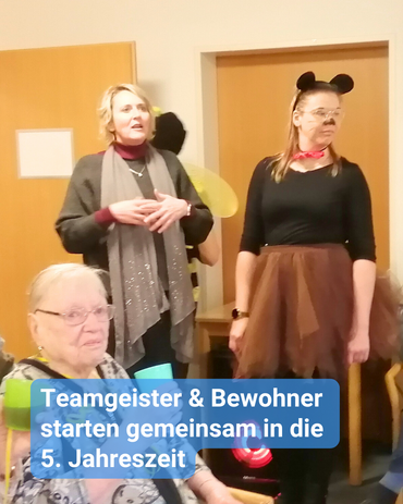 Im Vordergrund, eine Bewohnerin im Rollstuhl und im Hintergrund Mitarbeiterinnen, teilweise kostümiert. Bildbeschriftung: „Teamgeister & Bewohner starten gemeinsam in die 5. Jahreszeit“