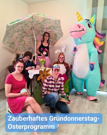 Blick auf Augenhöhe auf eine Gruppe von Menschen, hauptsächlich Frauen, die festliche Kleidung tragen und sich um einen kleinen Tisch unter einem blumengemusterten Sonnenschirm versammelt haben.  Ein großes, aufblasbares Einhornkostüm nimmt prominent rechts Platz. Die versammelten Personen befinden sich bei einer Osterveranstaltung, was durch die Dekorationen und Kostüme angezeigt wird; einige tragen Hasenohren. Die Szenerie spielt sich in einem Innenraum ab, dessen Wände und Böden in hellen Farben gehalten sind.  Am unteren Rand des Bildes befindet sich ein Text in Deutsch, der übersetzt lautet: "zauberhafter Gründonnerstag - Osterprogramm."