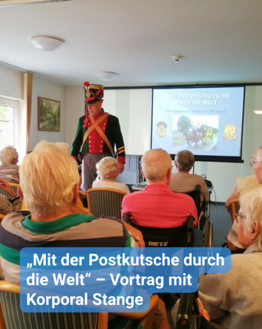 Eine Gruppe voller Senioren lauscht, dem Vortrag eines kostümierten Redners. Er trägt eine rot-schwarze Uniform. Bildunterschrift: „Mit der Postkutsche durch die Welt“ – Vortrag mit Korporal Stange