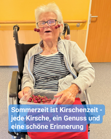 Seniorin im Rollstuhl hält stolz die Kirschernte in einer Schale auf dem Schoß fest. Sie lächelt fröhlich in die Kamera. Kirschen hängen dekorativ an ihrer Brille herab. Bildbeschriftung: Sommerzeit ist Kirschenzeit - jede Kirsche, ein Genuss und eine schöne Erinnerung