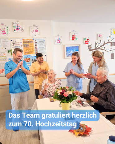 Am Tisch sitzt das Jubelpaar und hinter ihnen steht das Team der Pflege und Betreuung, und applaudiert. Bildbeschriftung: „Das Team gratuliert herzlich zum 70. Hochzeitstag.“