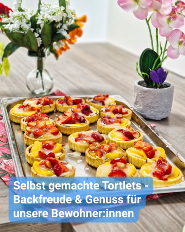 Ein Blech mit vielen kleinen, frisch belegten Obsttörtchen steht auf einem Tisch. Text: 'Selbst gemachte Tortlets - Backfreude & Genuss für unsere Bewohner:innen'.