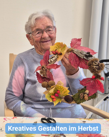 Eine Seniorin hält lächelnd einen selbst gebastelten Herbstkranz ins Bild. Bildbeschriftung: „Kreatives Gestalten im Herbst“