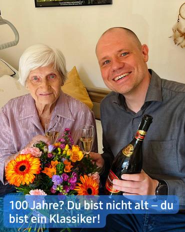 Nahaufnahme einer älteren Frau und eines jüngeren Mannes.  Die Frau mit kurzen, grauen Haaren sitzt und trägt eine mauvefarbene Strickjacke. Sie hält einen Strauß leuchtend bunter Blumen und blickt direkt zu den Betrachtern.  Der Mann sitzt an ihrer Seite. Er hat kurzes, hellbraunes Haar und trägt ein graues Hemd. Er hält eine Flasche Sekt in der Hand und lächelt breit. Sein Blick ist auf die Kamera gerichtet.  Beide sind in einem Raum positioniert, der wie ein Wohnzimmer oder ein Schlafzimmer wirkt. Im Hintergrund ist ein Teil eines Bettes oder Krankenbettes sichtbar. An der Wand hinter ihnen hängt ein Bild sowie ein Objekt, das wie ein Traumfänger oder Ähnliches aussieht, rechts und leicht hinter dem Mann.  Am unteren Rand ist eine feierliche Botschaft: "100 Jahre? Du bist nicht alt – du bist ein Klassiker!"