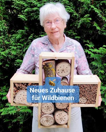 Eine Seniorin steht draußen vor einer grünen Hecke und hält ein großes Insektenhotel in den Händen. Bildbeschriftung: „Neues Zuhause für Wildbienen“