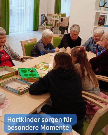 In mittlerer Entfernung zeigt sich eine Gruppe von Senioren und Kindern, die um einen Tisch herumsitzen und Brettspiele spielen. Der Schauplatz scheint ein Gemeindezentrum oder eine Senioreneinrichtung zu sein.  Die Erwachsenen sind auf Stühlen um einen hellen Holztisch versammelt, während die Kinder zwischen ihnen sitzen und näher zur Kamera ausgerichtet sind. Die Umgebung strahlt Helligkeit aus, mit hellen Wänden und grünen Vorhängen im Hintergrund, was auf eine warme und einladende Atmosphäre hinweist. Im Hintergrund sind rechts Bücherregale sichtbar und links scheint ein großes Fenster zu sein.  Eine Bildunterschrift am unteren Rand der Aufnahme lautet: "Hortkinder sorgen für besondere Momente."