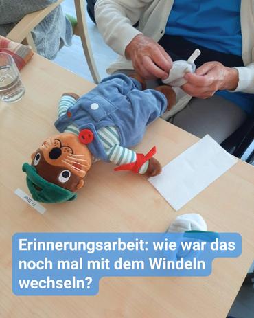 Eine Person sitzt an einem Tisch und wechselt die Windel eines Stoffotters. Der Otter liegt auf dem Rücken auf dem holzfarbenen Tisch, trägt eine blaue Latzhose und eine grüne Kappe. Die wechselnde Person trägt eine weiße Strickjacke über einem blauen Hemd. Auf dem Tisch sind ein Wasserglas, eine Serviette und ein Zettel sichtbar. Der Text auf dem Bild lautet: „Erinnerungsarbeit: wie war das noch mal mit dem Windeln wechseln?“