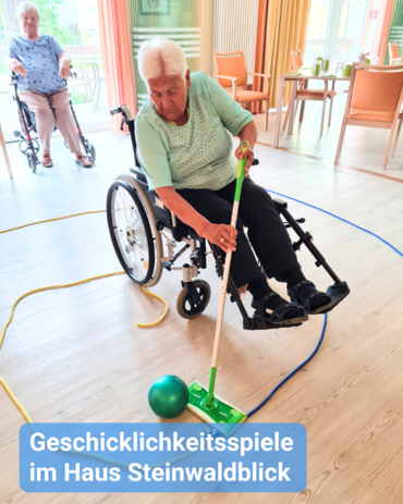 Eine ältere Frau im Rollstuhl spielt ein Geschicklichkeitsspiel, indem sie einen Ball mit einem Wischmob entlang eines Parcours bewegen muss. Im Hintergrund ist eine weitere ältere Dame im Rollstuhl zu sehen, die das Spiel beobachtet. Bildbeschriftung: Geschicklichkeitsspiele im Haus Steinwaldblick