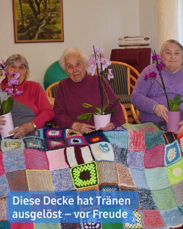 Drei ältere Damen sitzen auf Stühlen vor einer bunten, gehäkelten Decke.  Die Frauen lächeln sanft, jede hält ein Pflanzengefäß mit einer rosafarbenen Phalaenopsis-Orchidee. Die Dame in der Mitte trägt einen kastanienbraunen Pullover, während die beiden Frauen zu beiden Seiten Pullover in Lila- und Rosatönen tragen. Sie scheinen sich in einem behaglichen, häuslichen Ambiente zu befinden, möglicherweise in einem Pflegeheim oder einer Seniorenresidenz. Hinter ihnen hängt ein Gemälde einer Landschaft, und im Hintergrund erkennt man weitere Möbelstücke.  Die Decke im Vordergrund besteht aus zahlreichen Quadraten in verschiedenen Farben und Texturen, was auf ein handgefertigtes Patchwork-Design hindeutet. Die Bildunterschrift am unteren Rand des Bildes lautet: "Diese Decke hat Tränen ausgelöst - vor Freude."